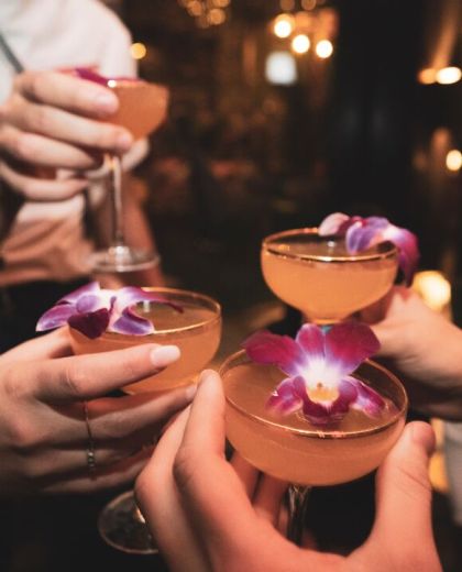四隻裝著橙色雞尾酒的金邊酒杯正在碰杯，酒中有紫色的可食用花朵