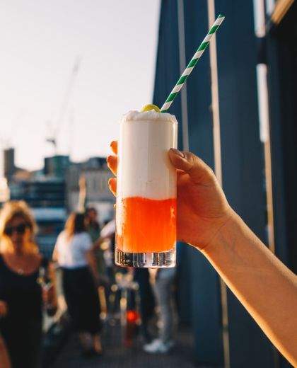 阳光下的citizenM伦敦塔酒店cloudM屋顶酒吧，擎着一杯带吸管和柠檬片装饰的橙白色饮料的手臂