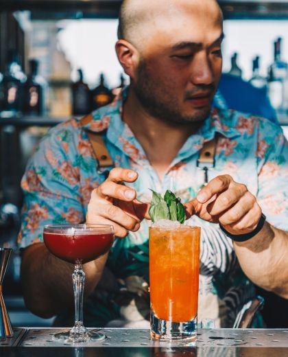身穿夏威夷衬衫的调酒师正在用薄荷叶装饰一杯橙色高杯鸡尾酒