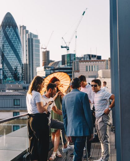 Group of men and women in casual clothes chat outside at cloudM rooftop bar with view of The Gherkin